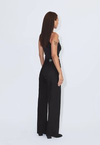 Frau mit langen braunen Haaren, die ein schwarzes ärmelloses Oberteil und weite schwarze Hosen trägt, seitlich vor einem schlichten weißen Hintergrund steht.
