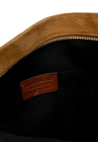 Interno di una borsa in suede marrone con rivestimento in tessuto nero. Presenta un'etichetta in pelle con la dicitura "Pelle Vera. Fabbricato in Italia." Dettaglio della cerniera in oro.