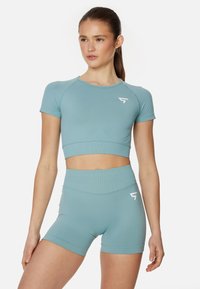 Svetlomodrý športový crop top s krátkymi rukávmi a zodpovedajúcimi šortkami s vysokým pásom. Obsahuje biely logo na hrudi a na boku. Hladká textura.