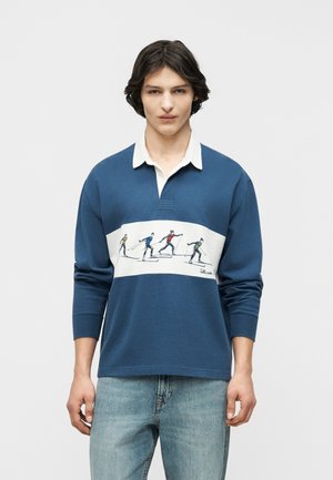 Mladý muž v modrej dlhorukávovej rugby košeli s bielym golierom a grafikou štyroch bežcov na lyžiach vpredu, v kombinácii so svetlými džínsami.