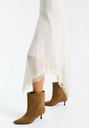 Frau trägt braune Wildleder-Stiefeletten mit spitzen Zehen und Katzenabsätzen, kombiniert mit einem weißen, transparenten Rock mit Spitzenbesatz.