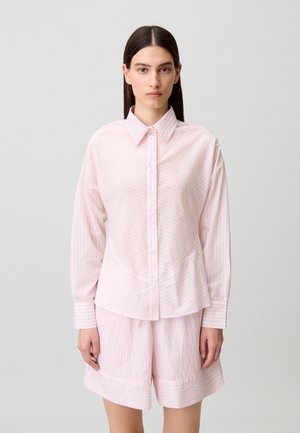 Femme aux cheveux raides et foncés portant une chemise à boutons rose pâle et un short ample assorti, posant devant un fond clair uni.