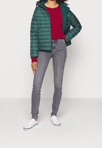 Femme portant une doudoune verte à pois par-dessus un pull rouge, un jean skinny gris et des baskets blanches, debout devant un fond uni.