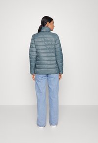 Casaco acolchoado em teal com gola alta e textura matelassada. Mangas longas e vista traseira, combinado com jeans de perna larga em azul claro.