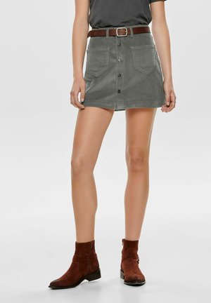 Femme portant une jupe mini en velours corduroy gris avec une ceinture en cuir marron et des bottines en daim marron sur fond blanc.
