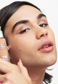 Foundationflaska i klart glas med en beige vätska, hålls nära ett ansikte med medeltonad hy, med ljusa ögonfransar och en mjuk läppfärg.