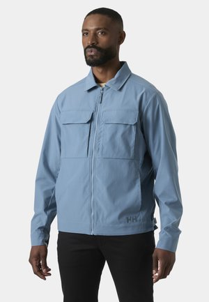 Mann trägt eine hellblaue Jacke mit Reißverschluss und zwei Brusttaschen sowie schwarze Hose, steht vor einem schlichten grauen Hintergrund.