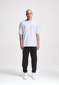 Weißes Baumwoll-T-Shirt mit lockerer Passform und kurzen Ärmeln, kombiniert mit schwarzen Jogginghosen und weißen Sneakers, vor einem schlichten Hintergrund stehend.