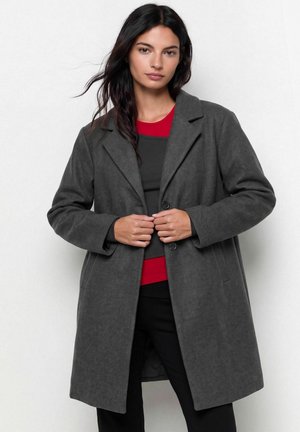 Une femme porte un long manteau gris foncé sur un haut noir et rouge et le tient fermé devant un fond clair uni.