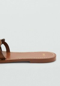 Brown leather sandals with crossed straps, flat sole, and decorative stitching along the edge. Gold logo printed on the footbed.
