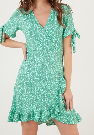 Green floral wrap dress with short sleeves tied at elbows and ruffled hem worn by person with hand on hip.