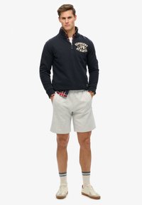 Pullover in pile blu navy con zip a tre quarti e logo "Superdry Japan Athletic", pantaloni corti grigi, sneakers color crema e calze a righe. Il modello sta in piedi con sicurezza.