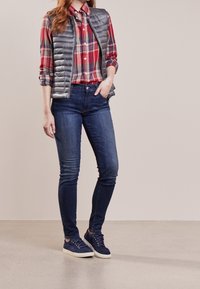 Šedá péřová vesta přes červenou károvanou košili, spárovaná s tmavě modrými skinny džíny a námořnickými teniskami. Hladké textury a přiléhavý styl.