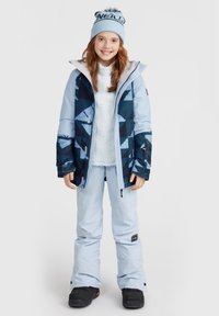 Veste de neige bleue avec des motifs géométriques, associée à un pantalon de neige bleu clair et des bottes de neige noires. La fille porte un bonnet bleu clair.