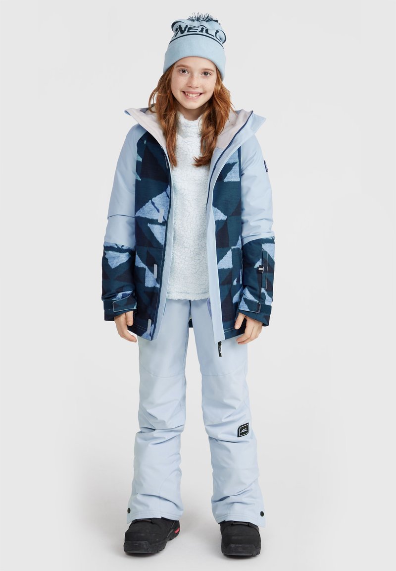 Veste de neige bleue avec des motifs géométriques, associée à un pantalon de neige bleu clair et des bottes de neige noires. La fille porte un bonnet bleu clair.