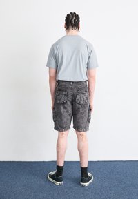 Vaaleanharmaa t-paita, mustat farkku cargo-shortsit, joissa on taskut, ja tummat lenkkarit henkilöllä, joka näkyy selältä yksinkertaista taustaa vasten.