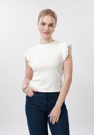 Jeune femme aux cheveux blonds portant un haut blanc sans manches à volants et un jean bleu foncé, debout avec une main dans la poche, souriant doucement.