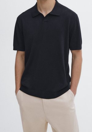 Man draagt een zwart poloshirt met korte mouwen en beige broek, handen in de zakken gestoken, tegen een effen achtergrond.