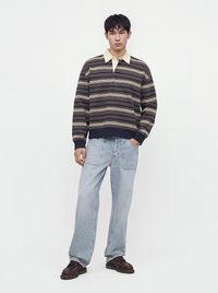 Jeune homme portant un polo à manches longues rayé, jean bleu clair avec de grandes poches avant et chaussures marron foncé, debout avec une main dans la poche.