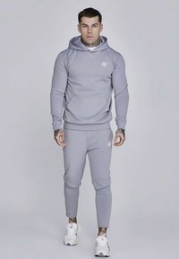 Svetlosivá športová mikina s kapucňou a klokankovým vreckom, doplnená o nohavice s zúženými nohavicami v rovnakom odtieni. Materiál pôsobí hladko a je s minimálnym počtom log. Biele tenisky.