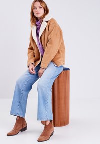 Giacca in suede marrone con colletto in shearling bianco, abbinata a un dolcevita viola, jeans azzurri chiari e stivaletti marroni.