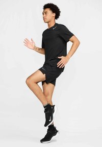 Camiseta atlética negra y pantalones cortos, ambos con un sutil logo de Nike. Llevado con zapatillas negras y calcetines grises, con movimiento de piernas visible.