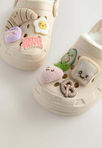 Sabots beige avec divers patchs colorés : un œuf souriant, un avocat, un bretzel, et la phrase "Meilleurs amis pour toujours", avec une doublure douce.