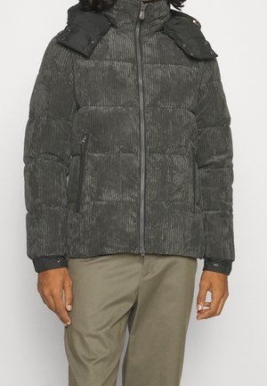 Homme portant une veste matelassée en velours côtelé gris foncé avec fermeture éclair devant et pantalon beige, debout devant un fond uni.