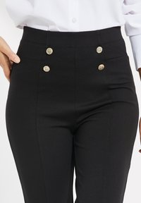Hoge zwarte broek met een gladde textuur en gouden knoopaccenten, met platte voorzakken en een getailleerde pasvorm.