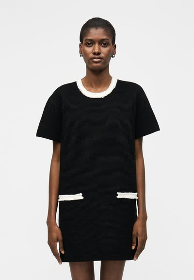 Femme portant une robe noire texturée à manches courtes avec un bord à franges blanches au niveau de l'encolure et des poches, debout devant un fond uni.