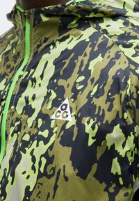 Κοντινό πλάνο ατόμου που φοράει μπουφάν παραλλαγής σε πράσινο και μαύρο με φωτεινό πράσινο φερμουάρ και λευκό λογότυπο ACG στο στήθος.