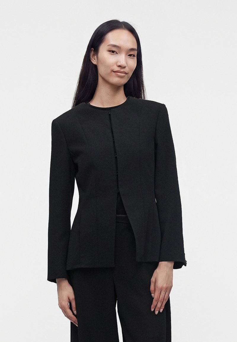 Femme aux cheveux lisses et foncés debout devant un fond uni, portant une veste noire cintrée et un pantalon noir assorti, de face.