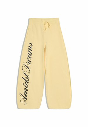 Lichtgele broek met wijde pijpen, voorzakken en een taille met trekkoord, met zwarte cursieve tekst "Amidst Dreams" langs het linkerbeen.