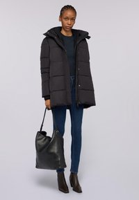 Černá péřová bunda s kapucí, prošívaný design, patentky, nošená přes námořnickou modrou top. Spárována se skinny džíny a velkou černou taškou.