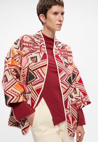 Többszínű mintás kardigán geometrikus formákkal piros, narancssárga és fekete színekben; puha szőtt textúrával és nyitott elöl, semleges szegéllyel.