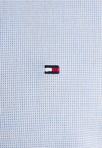 Tissu texturé bleu clair et blanc avec un petit logo rectangulaire comportant des bandes horizontales noires, blanches et rouges au centre.
