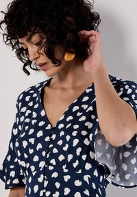 Marineblaues Kleid mit weißem Herzmuster, V-Ausschnitt, kurzen, ausgestellt Ärmel und Knopfdetails. Modell mit lockigem Haar trägt orangefarbene Ohrringe.