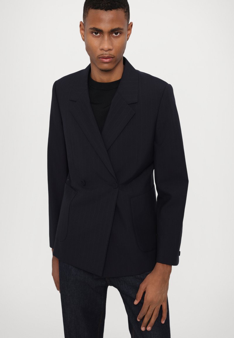 Marineblauer Nadelstreifen-Blazer mit einem doppelt knöpfbaren Design, Reverskragen, zwei Fronttaschen und passgenauer Schnittführung. Der Stoff wirkt angenehm glatt.