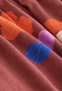 Tekstil med en rustfarvet baggrund, prydet med levende, abstrakte former i orange, lilla og blå. Blødt, tekstureret stof med lagdelte folder.