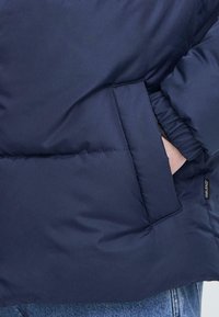 Jachetă puffer bleu marin, cu un buzunar mare cusut și manșete elasticizate, fabricată dintr-un material moale și neted. Eticheta brandului este vizibilă.