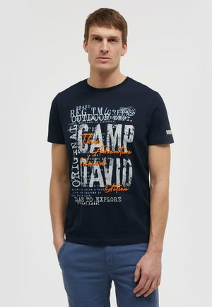 Mann trägt ein dunkelblaues T-Shirt mit weißem und orangefarbenem Text und steht mit einer Hand in der Tasche seiner blauen Hose vor einem schlichten Hintergrund.