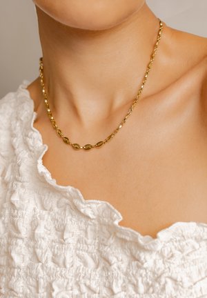 Goldene Kettenhalskette mit länglichen ovalen Gliedern und kleinen runden Akzenten, getragen über einer weißen strukturierten Bluse mit Rüschekanten.
