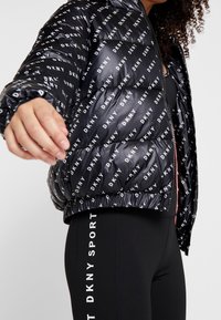 Černá péřová bunda s opakujícím se bílým logem "DKNY", s předním zipem, elastickými manžetami a lemem. Nošena s černými legínami s textem "DKNY SPORT".