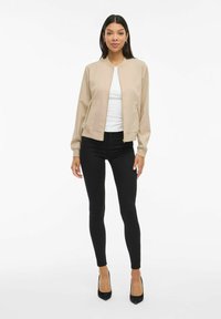 Femme portant une veste bomber beige, une chemise blanche, un pantalon skinny noir et des talons noirs, se tenant devant un fond blanc.