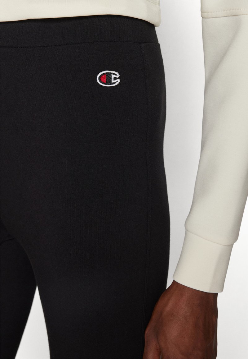 Leggings neri in cotone con un design aderente, caratterizzati da un piccolo logo ricamato rosso e bianco sul lato e una texture liscia.