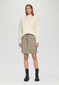 Vrouw in een crèmekleurige chunky gebreide trui, beige corduroy rok met trekkoord en zwarte leren laarzen tot halverwege de kuit, staand tegen een effen achtergrond.