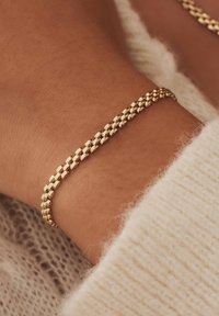 Goldfarbenes Metallarmband mit strukturiertem Design und ineinandergreifenden Gliedern, getragen an einem Handgelenk neben einem weichen, cremefarbenen Pullover.