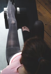 Black foam roller on a dark mat. A leg in black tights and a bare foot rests on the roller. Sunlight highlights the smooth textures.