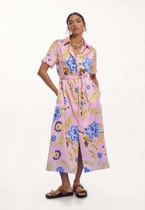 Mujer que lleva un vestido midi rosa con estampado floral en azul y amarillo, un cinturón en la cintura, joyas de oro y sandalias marrones con tachuelas, de pie con las manos en los bolsillos.