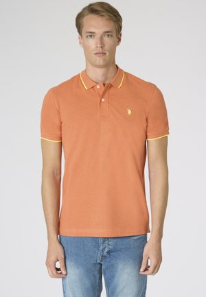 Uomo in piedi di fronte, indossa una polo arancione con rifiniture gialle e jeans azzurri chiari su uno sfondo semplice.